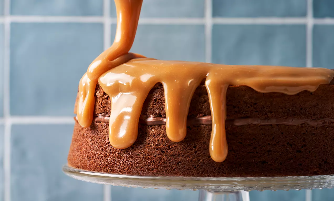 Twee-laagse chocoladecake bedekt met dikke, glanzende karamelsaus die langzaam naar beneden druppelt, gepresenteerd op een glazen taartstandaard