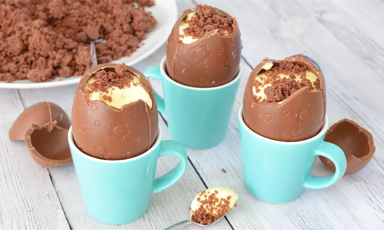 Chocolade paaseieren gevuld met gele kwarktaartcrème en bruine kruimels, rechtopstaand in lichtblauwe mokken op een houten tafel