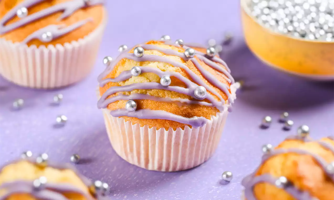 Zachte goudbruine muffins met paarse glazuurstrepen en zilverkleurige parels in witte vormpjes op een lila ondergrond