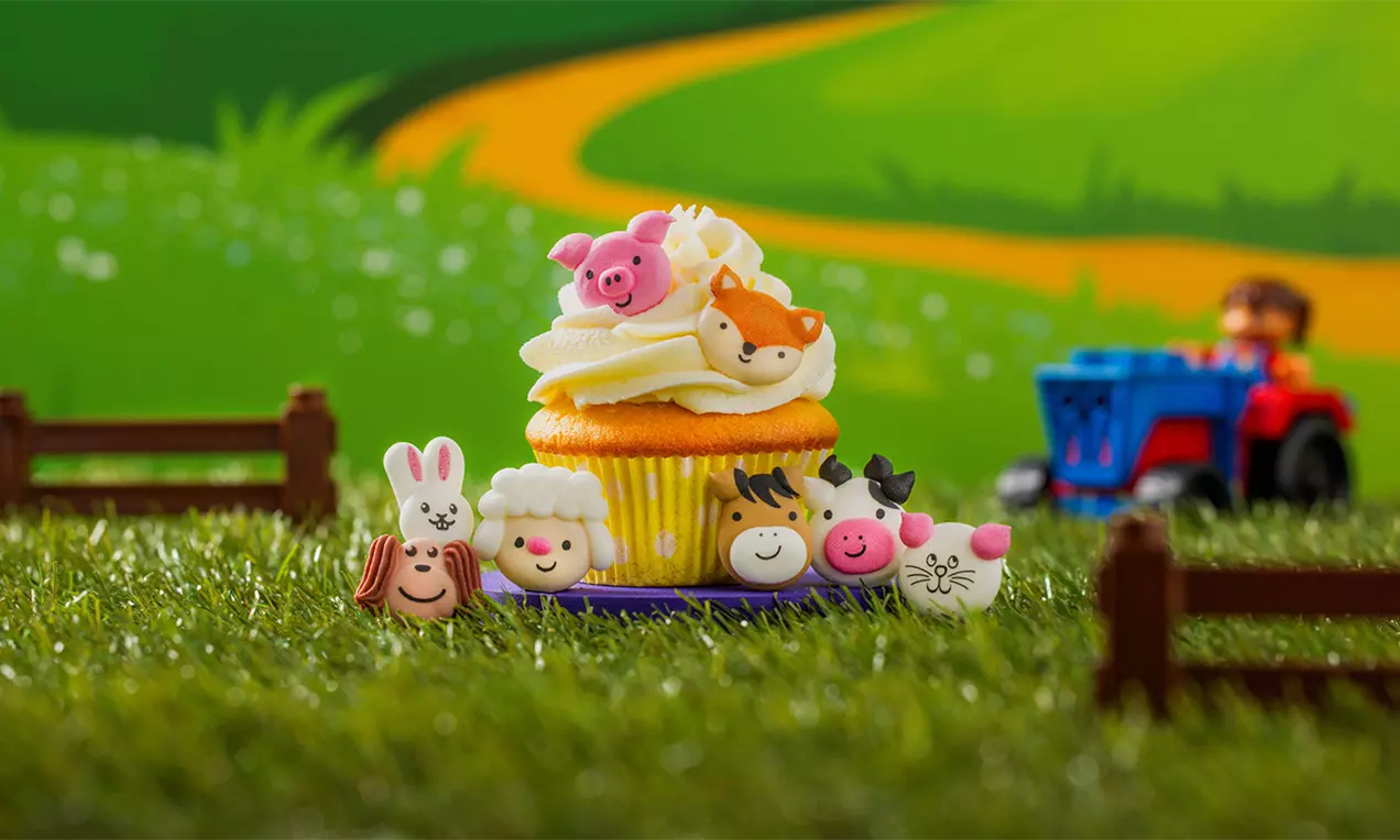 Cupcake met gele cake en witte room, versierd met kleurrijke boerderijdiertjes in een groene grasdecoratie met een speelgoedtractor