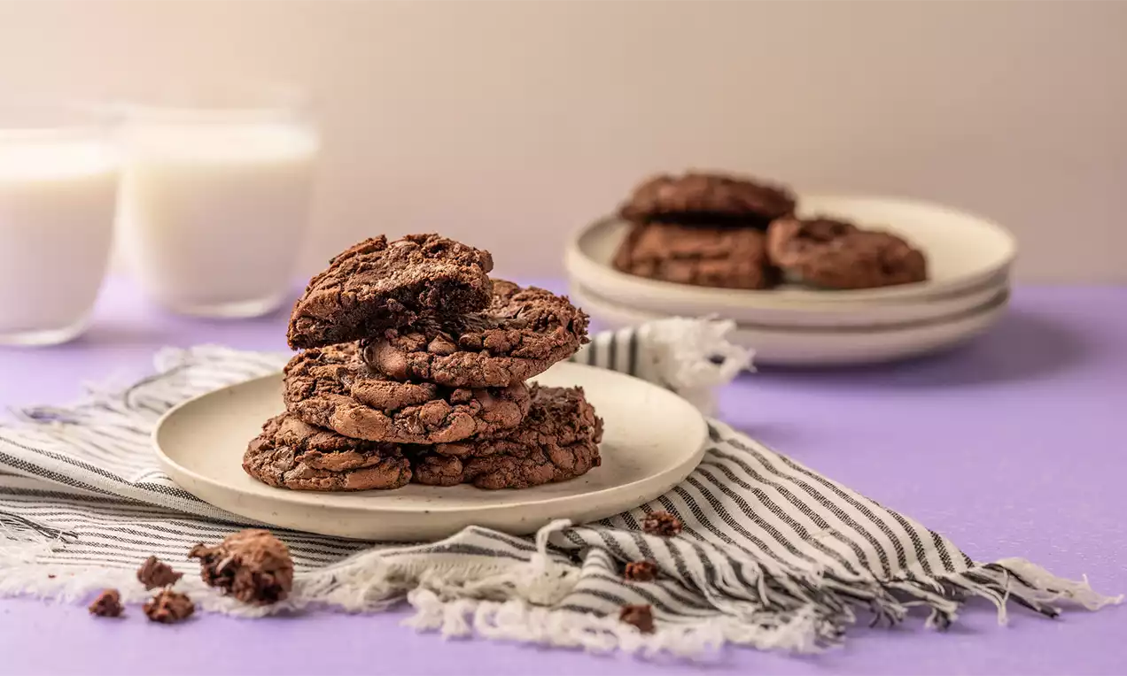 Donkerbruine, gebarsten brownie cookies op een wit bordje met een gestreepte doek, met glazen melk en borden op een paars oppervlak