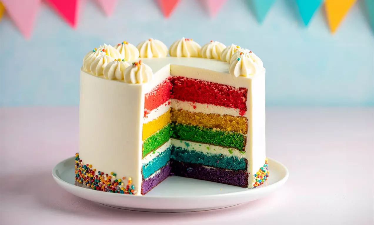 Regenboogtaart met zes gekleurde lagen en witte crème, versierd met toefjes slagroom en regenboogsprinkles op een wit bord, AI-labeled image