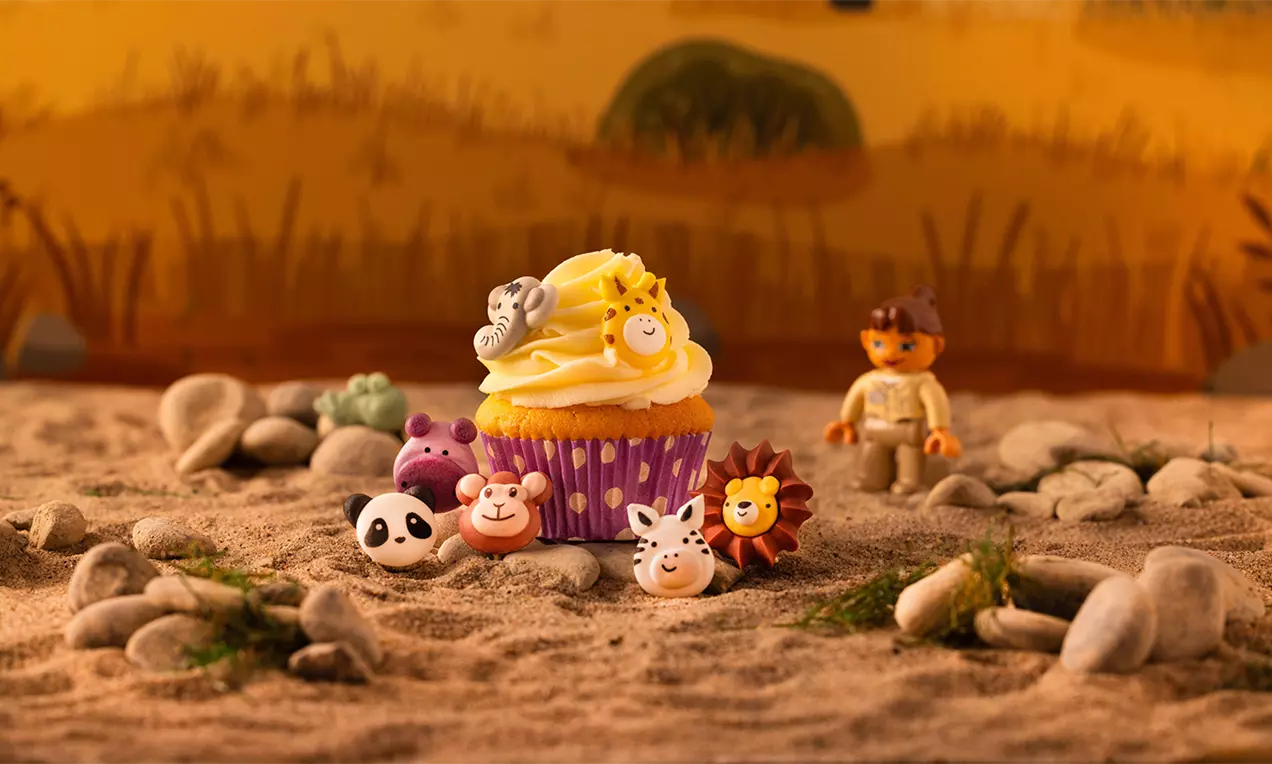 Cupcake met lichtgele roomtoef en safaridiertjes van fondant in levendige kleuren op een zandig decor met stenen en grasplukjes