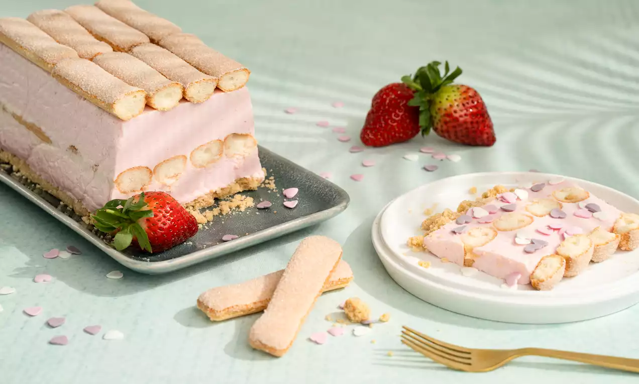 Roze aardbei-kwarktaart-tiramisu met lagen lange vingers op een rechthoekige schaal en een bordje, versierd met schijfjes aardbei en hartjesconfetti