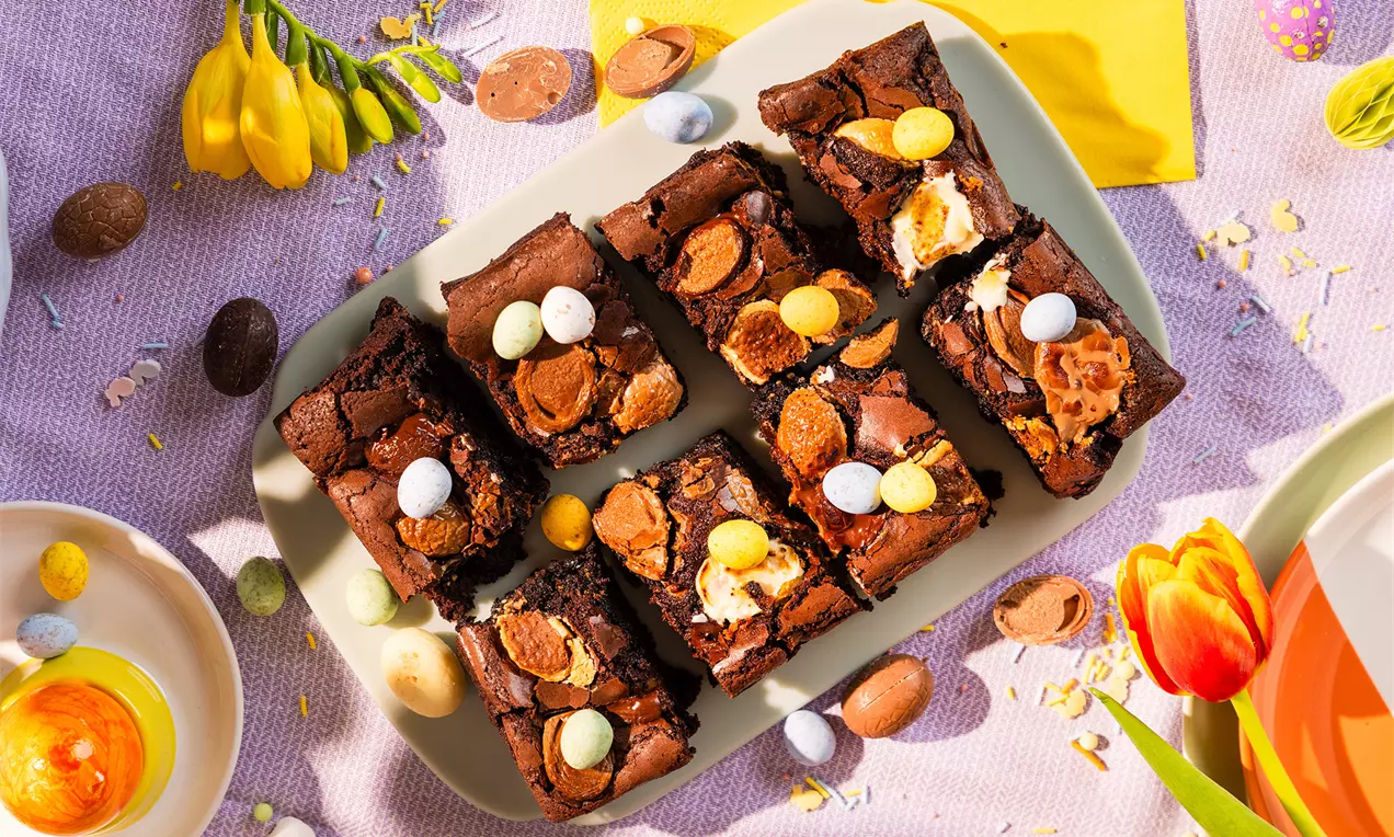 Donkerbruine paasbrownies met een smeuïge textuur, stukjes chocolade en gekleurde paaseitjes, gerangschikt op een lichtgrijze schaal