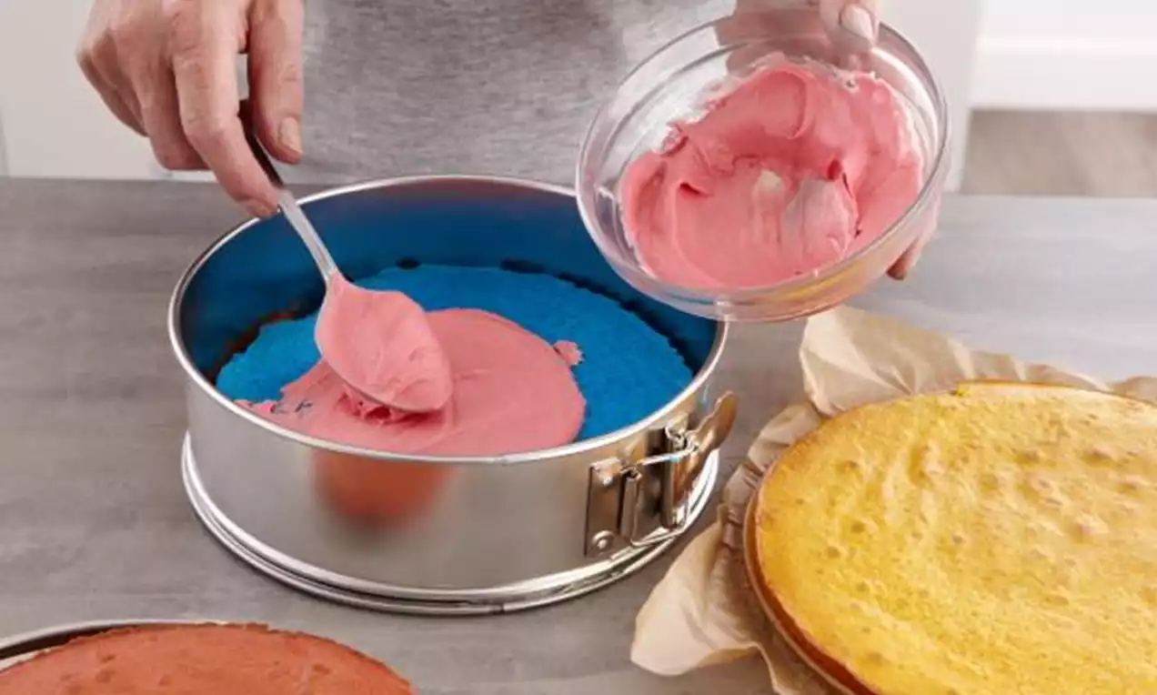 Een springvorm met blauwe cake en een bovenlaag van roze glazuur die met een lepel wordt aangebracht, naast een gele en rode cake op bakpapier