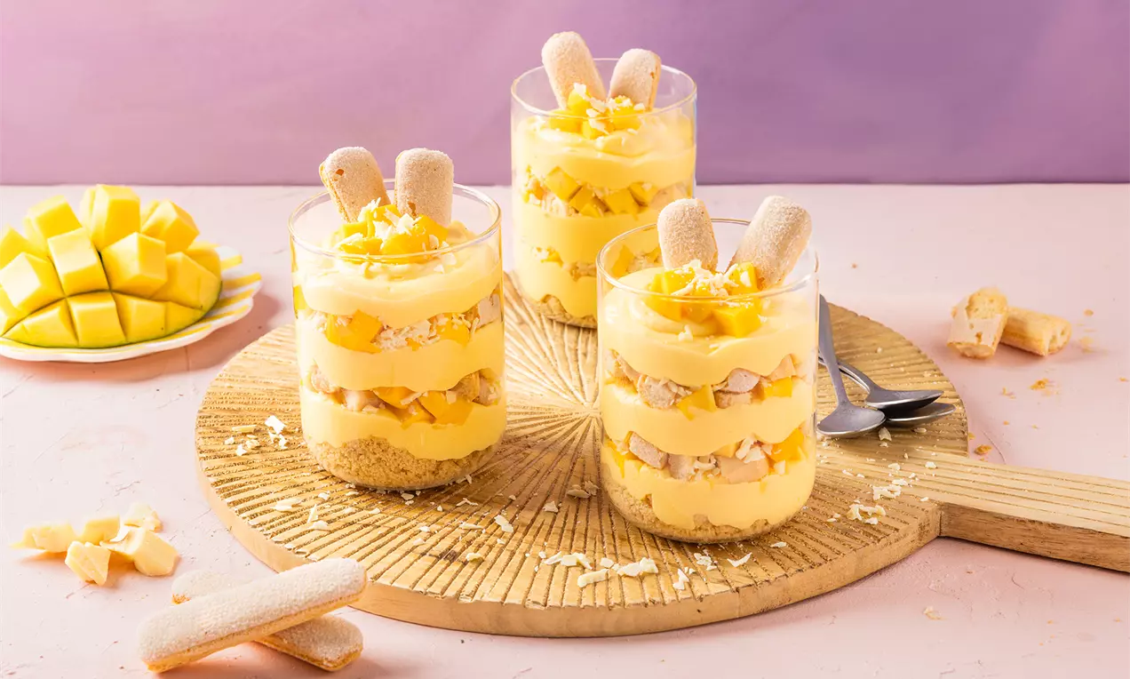 Mangotiramisu met laagjes geel mangocrème, lange vingers en room, gegarneerd met mangostukjes en witte chocoladerasp in glazen op een houten bord