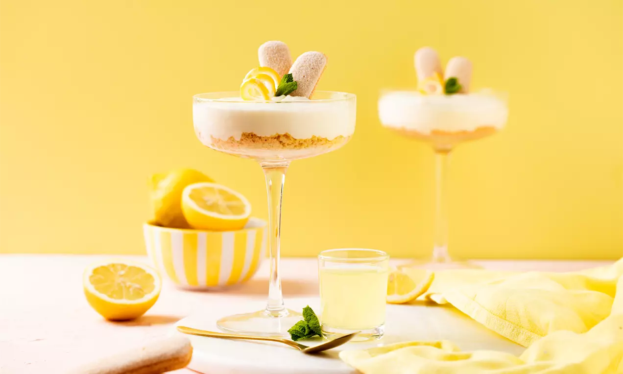 Limoncello tiramisu in een hoog glas met romige lagen, een kruimelbodem, lange vingers, citroenzeste en muntblaadjes, geserveerd op een lichtgele achtergrond